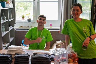 Zwei Jugendliche an einem Tisch, auf dem T-Shirts, Wasserflaschen und Äpfel liegen.