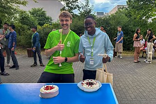 Zwei Jugendliche an einem Tisch, auf dem zwei Torten mit kleinen Kerzen stehen.