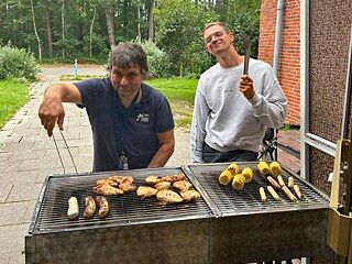 Zwei Männer stehen am Grill, auf dem Maiskolben, Hähnchenbrust und Würstchen liegen.