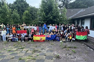 Eine große Gruppe Jugendlicher stehen vor einem Gebäude, sie halten Flaggen und lächeln in die Kamera.