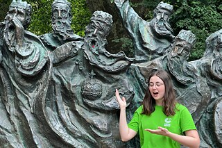 Junge Frau parodiert vor der Skulpturgruppe „Fährmann hol’ über“ in Speyer