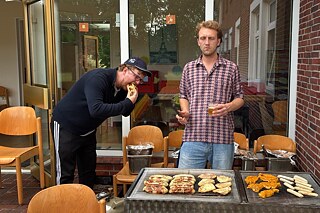 Ein junger Mann steht am Grill, ein anderer beißt herzhaft in ein Brötchen.