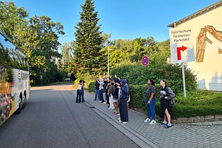 Einige Jugendliche stehen an einem Straßenrand und verabschieden sich. Links ist ein großer Bus zu sehen.