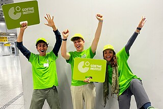 Drei junge Erwachsene mit Identifikationsschildern vom Goethe-Institut an einem Flughafen