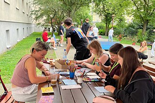 Jugendliche sitzen an Biertischen in einem großen Garten und malen.