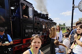 Jugendliche an einem Bahnhof, im Hintergrund ist eine Dampflokomotive zu sehen.