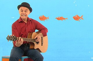 Ein Mann spielt Gitarre, im Hintergrund eine blaue Wand mit drei orangefarbenen Fischen und Luftblasen.