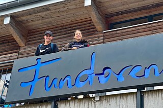 Zwei junge Menschen winken von einem Balkon, daran befestigt der Schriftzug „Fundorena“.