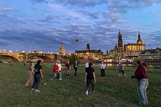 Postkarten Dresden Jugendliche spielen Volleyball auf einer Wiese, im Hintergrund die hell beleuchtete Siluette von Dresdens Altstadt.