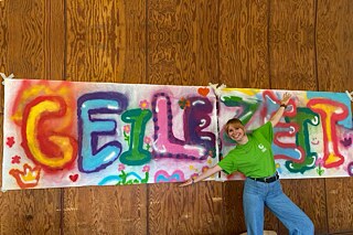 Eine junge Frau steht vor einem Graffiti-Schriftzug auf dem steht in bunten Lettern: „Geile Zeit“