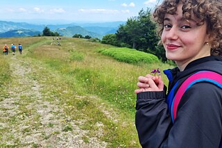 Eine junge Frau auf einem Wanderweg hält eine kleine Blume in der Hand und lächelt. Im Hintergrund sind Hügel und Bäume zu sehen.