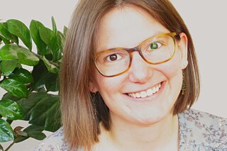 Frau mit Brille lächelt in die Kamera, im Hintergrund eine Zimmerpflanze