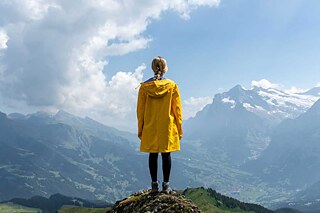 Frau in gelbem Regenmantel steht auf einem Felsen und schaut auf eine Landschaft