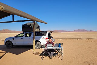 Ein Mann sitzt an einem Klapptisch, vor ihm stehen zwei Laptops. Im Hintergrund ein Geländewagen mit Zelt auf dem Dach und eine Wüstenlandschaft.