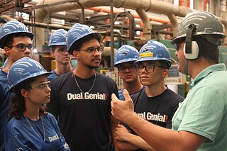 Sechs Jugendliche mit blauen Schutzhelmen und T-Shirts mit der Aufschrift „Dual-Genial“ hören einem Erwachsenen interessiert zu.