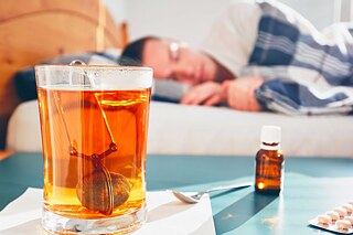 Eine männliche Person liegt im Bett, im Vordergrund ein Tee und ein Blister mit Tabletten