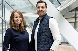 Ein Mann und eine Frau stehen nebeneinander vor einer Fensterfront und lächeln