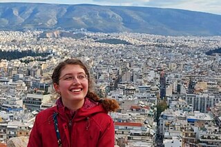 Frau in roter Jacke vor einem Stadtpanorama, im Hintergrund Berge