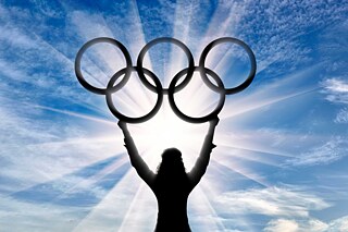 Die Siluette einer Athletin (von hinten zu sehen) hält die olympischen Ringe in den Himmel.
