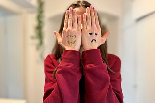 Eine junge Frau verdeckt ihr Gesicht mit den Händen. Auf den Handrücken sind links ein lächelndes Gesicht, rechts ein trauriges Gesicht gemalt.
