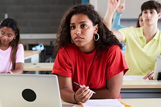 Schülerin sitzt an einem Tisch, vor ihr ein Laptop und Schreibutensilien. Im Hintergrund sitzen drei weitere Personen, von denen sich eine meldet.