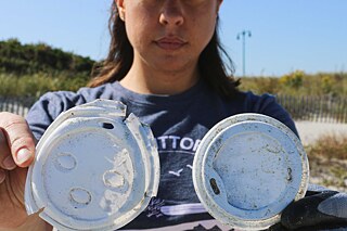 Person steht am Strand in einem Pullover und hält zwei Kaffeebecherdeckel in die Kamera