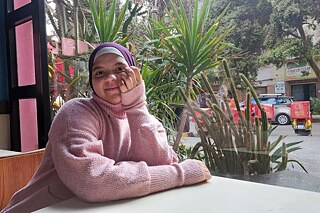 Eine Jugendliche mit Hidschab sitzt in einem Café an einem Tisch. Im Hintergrund ist eine Straße zu sehen, auf der rote Liefermopets stehen.