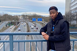 Junger Mann steht auf einer Autobahnbrücke. Im Hintergrund kann man ein blaues Autobahnschild erkennen.