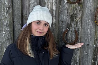 Eine junge Frau warm gekleidet mit heller Strickmütze steht vor einer Holzwand an der Hufeisen mit der Öffnung nach oben genagelt sind.