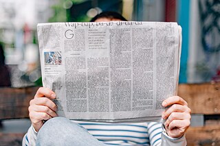 Mensch sitzt auf einer improvisierten Bank und liest in einer urbanen Umgebung Zeitung.