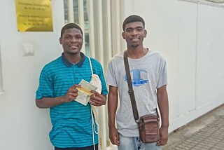 Zwei Personen stehen nebeneinander vor dem Gebäude des Konsulats von Mosambik. Die linke Person trägt ein blau gestreiftes Poloshirt und hält ein Buch mit dem Titel ‚MOZAMBIQUE‘. Die rechte Person trägt ein graues T-Shirt mit blauem Logo und eine braune Umhängetasche. Im Hintergrund ist eine goldene Plakette mit der Aufschrift ‚Mozambique Consulate‘ sichtbar.