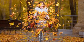 Eine junge Radfahrerin fährt durch umherwirbeldes, bunt gefärbtes Herbstlaub.