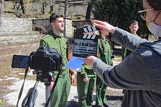 Schülerin hält Filmklappe vor Videokamera, drei Schüler in Uniformen stehen in einer Reihe
