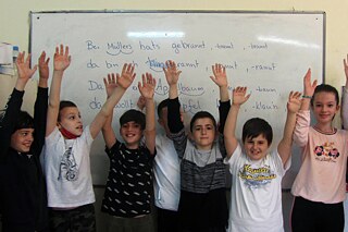 Schüler und Schülerinnen stehen vor einem Whiteboard und halten alle beide Hände in die Luft
