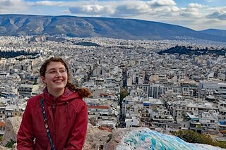 Frau in roter Jacke vor einem Stadtpanorama, im Hintergrund Berge