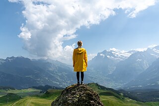 Frau in gelbem Regenmantel steht auf einem Felsen und schaut auf eine Landschaft