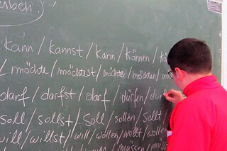 Schüler mit roter Jacke schreibt an der Tafel Verben: können, müssen, sollen