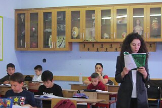 Schüler und Schülerinnen sitzen in einem Klassenzimmer an ihren Tischen und lesen, Lehrkraft läuft durch den Raum