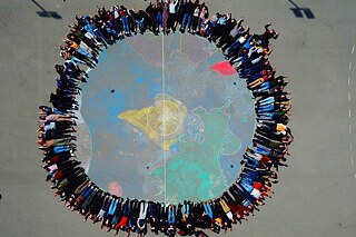 Luftaufnahme einer auf den Boden gemalten Weltkugel, Schülerinnen und Schüler liegen im Kreis außenherum