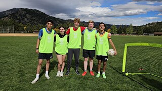 Fünf junge Menschen in gelben Fußballtrikots stehen vor einer bergigen Landschaft. Einer von ihnen hält einen Ball.