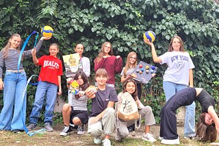 Eine Gruppe von Personen steht vor einer grünen Hecke und hält verschiedene Sport- und Spielgeräte, darunter Volleybälle, ein Springseil und ein Brettspiel.