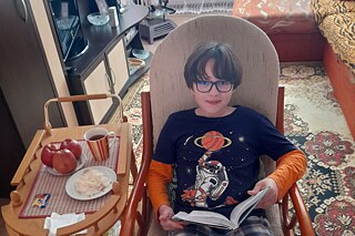 Eine Person sitzt in einem gepolsterten Stuhl und liest ein Buch. Daneben steht ein kleiner Tisch mit einer Tasse, zwei Äpfeln und einem Teller mit Essen.