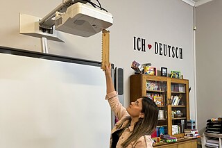 Eine Person steht in einem Klassenraum und nutzt ein langes Holzlineal, um einen Projektor über einer Tafel zu bedienen.