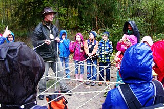 Kinder und Erwachsene in winterlicher Kleidung stehen im Wald und halten ein kreuz und quer gespanntes Seil.