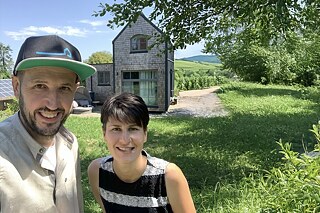 Zwei junge Menschen stehtn auf einer Wiese, im Hintergrund sieht man ein kleines, mit Schindeln verkleidetes Haus.