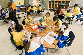 Eine Schülergruppe und Lehrerinnen/Erzieherinnen sitzen an Tischen in einem Klassenzimmer.