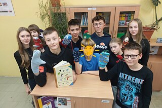 Kindergruppe in einem Klassenraum zeigt bunte Sockenpuppen, ein Buch liegt auf einem kleinen Schrank im Vordergrund.