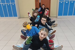 Kinder sitzen in einer Reihe auf dem Boden, zeigen bunte Sockenpuppen, vorne liegt ein Buch. Im Hintergrund sind blaue Schließfächer und eine Tür.