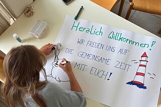 Eine junge Frau mit langen, blonden Haaren sitzt an einem Tisch und malt ein Plakat. Darauf steht „Herzlich Willkommen! Wir freuen uns auf die gemeinsame Zeit mit Euch“