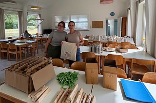 In einem großen Raum stehen zwei junge Frauen hinter einem  großen Tisch; die eine hält eine Stofftasche in der Hand. Auf dem Tisch stehen Kartons, daneben liegen Holzstäbe, eine blaue Mappe und grüne Schlüsselbänder.